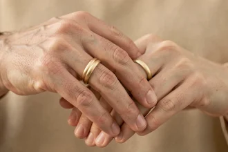 Deux mains adultes enlacées portant des bagues duo homme femme en or sur fond neutre chaleureux