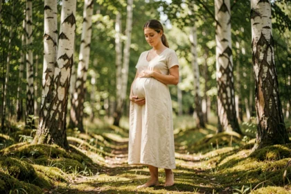 Femme enceinte en robe ivoire dans une forêt de bouleaux, tenant son ventre avec sérénité, photo de grossesse naturelle en plein air